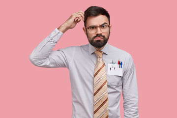 Hesitant unsure male designer scratches head and looks with serious expression, thinks about new sketh, wears formal shirt, has pens and pencil in pocket, models over pink background, finds decision
