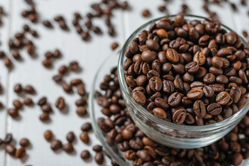 Glass bowl with saucer filled with coffee on background of scattered grains on white table.