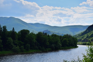 Silver river among the forests. Illuminated by sunlight mountains in the distance, a thick dark forest stands on the river bank. Blue sky with clouds. River flowing between the mountains.