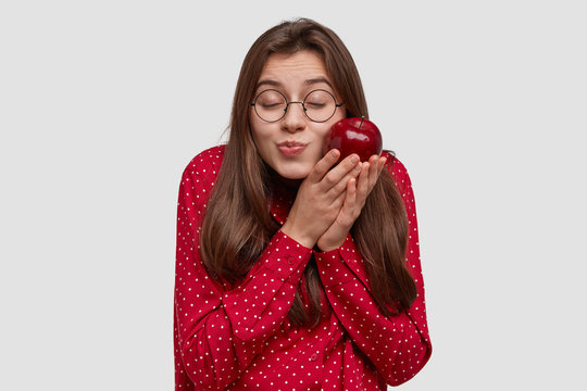 Pleased attractive woman with dark hair, keeps red apple near cheek, closes eyes, has pleasure, wears round glasses, wears polka dot shirt, isolated over white background. Female and dieting concept