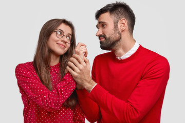 Horizontal shot of pleased woman looks at husband who has pleading expression and holds her hand, wear red clothes, have pleasant talk, isolated over white background. People, love, relations
