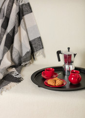 Round black metal tray with a coffee maker and a cup on a white sofa with a gray plaid and pillows.