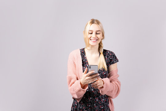 Studio shot of gorgeous young blonde woman with long straight hair wearing soft pink loose knit oversized sweater and feminine black dress. Gray isolated background, copy space, close up, portrait.