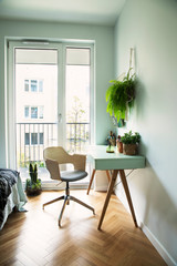 Modern scanidinavian interior of home office with design table, armchair, office accesories, big window and plants. Stylish and sunny space with wooden brown parquet. 