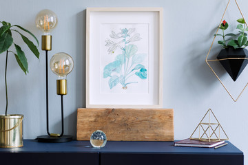 Interior floral poster mock up with vertical wooden frame, table lamp, avocado plant, accessories and hanging plants in geometric pot on the grey wall background. Concept with navy blue shelf. 