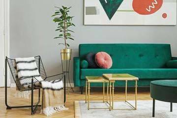 Luxury and modern home interior with green velvet design sofa, armchair, tables and pouf. Tropical plant in metal stand. Grey walls with abstract painting. Stylish decor of sitting room.
