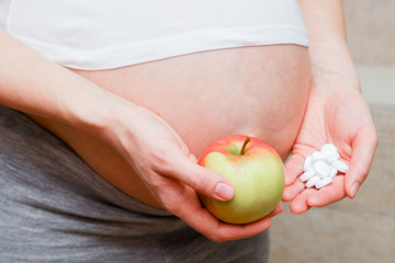 A pregnant girl in a T-shirt and holding a fresh apple and white pills. The concept of choosing a healthy diet or medicine.