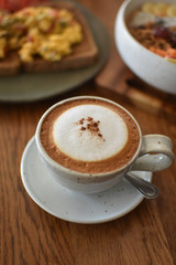 At stylish café in Thailand.Tasty cappuccino, with a hint of cinnamon in the middle of milk foam, looking from the side angle with delicious egg toast and fruits bowl in the background.Portrait mode.