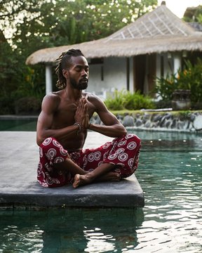 Skinny African American Male Wearing Indian Style Pants Sitting In Meditation Pose By The Swimming Pool