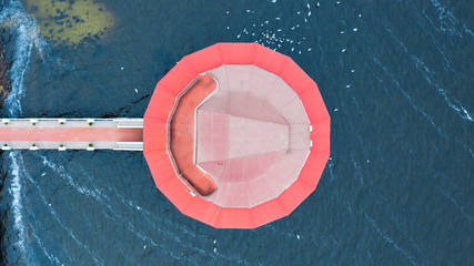 Aerial top view on blue water surface in a river around a round red-roofed building with a pier.