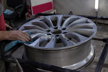 Master body repair man is working on preparing the surface of the aluminum wheel of the car for subsequent painting in the workshop, cleaning and leveling the wheel with the help of abrasive material