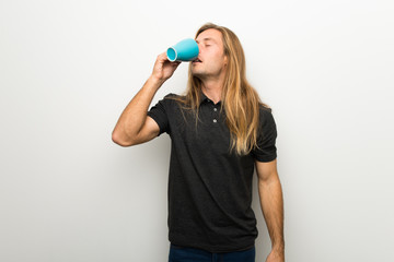Blond man with long hair over white wall holding a hot cup of coffee
