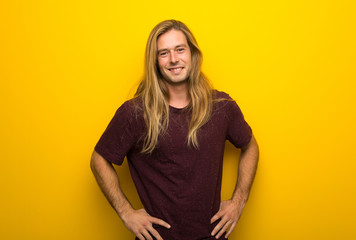 Blond man with long hair over yellow wall posing with arms at hip and laughing looking to the front
