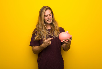 Blond man with long hair over yellow wall holding a piggybank