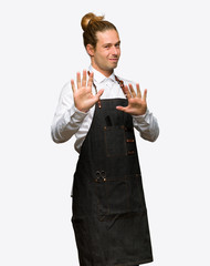Barber man in an apron is a little bit nervous and scared stretching hands to the front on isolated background
