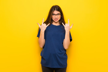 Young woman with glasses over yellow wall making rock gesture