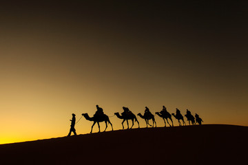 Fototapeta na wymiar Sillhouette of camel caravan with happy peopple going through the desert at sunset with red sky in background