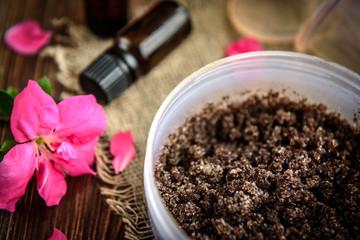 Coffee body scrub, sugar and coconut oil, essential oils, massage vacuum jars on dark wooden rustic table with pink flowers. Homemade cosmetic for peeling and spa care.