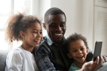 Happy african american dad laughing using smartphone with little son daughter at home, black daddy...