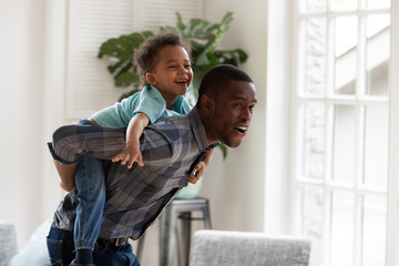 Happy african dad gives little funny son piggyback ride play at home, cheerful single black daddy carrying kid on back have fun together, child boy enjoy weekend activity with father in living room