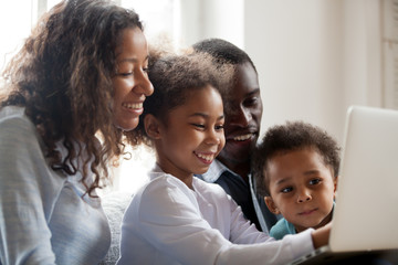 Happy black family with kids having fun using laptop application together at home, african parents and children laughing doing shopping, watching funny internet video, making online computer call