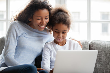 Smiling african american mother and kid daughter have fun shopping online at home, happy black mom teaching little mixed race child girl learn use laptop, watch cartoon, make video call on computer