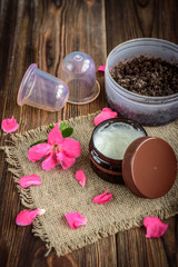 Coffee body scrub, sugar and coconut oil, essential oils, massage vacuum jars on dark wooden rustic table with pink flowers. Homemade cosmetic for peeling and spa care.