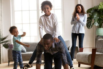 Happy african american dad riding mixed race daughter on back, playful black family enjoying playing funny game with kids together in living room, parents and children laughing having fun at home