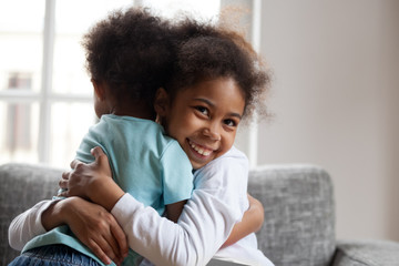 Smiling african girl sister embracing little boy brother at home, mixed race preschool kids hugging cuddling, happy black children having fun together, 2 siblings friendship good relations concept