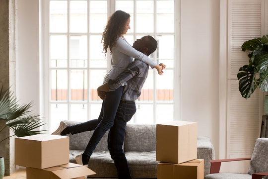 Happy African American First Time Home Buyers Couple Celebrating Relocation Or Buying House In Living Room With Boxes, Excited Black Man Lifting Embracing Woman Enjoying Moving Day, Family Goals