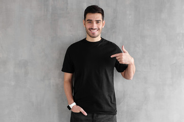 Young handsome man isolated on gray textured wall, smiling while pointing with index finger to black t-shirt, copyspace for advertising - obrazy, fototapety, plakaty