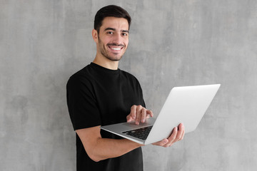 Portrait of young man in black t-shirt standing against textured wall, holding laptop and watching media with happy smile, sharing web content