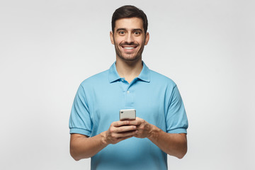 Young man standing isolated on gray background, holding smartphone, looking at camera and smiling nicely