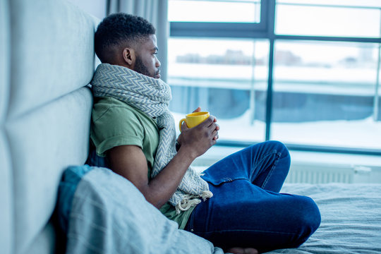 Gloomy african student has to stay home and to miss important classes at college because he has caught a season cold. Upset guy in warm scarf holding a cup with liquid medicine and and looking aside