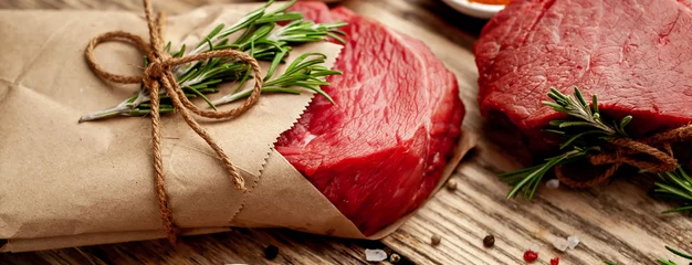 Fotobehang two pieces of meat from the butcher shop, one piece of meat wrapped in paper. A piece of beef in the form of a steak on a wooden background with ingredients © александр таланцев