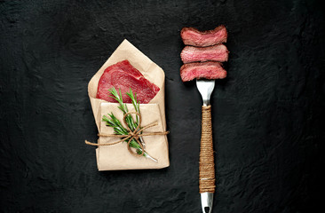 beef steak on a fork, next to a piece of raw meat in wrapped paper on a dark concrete background