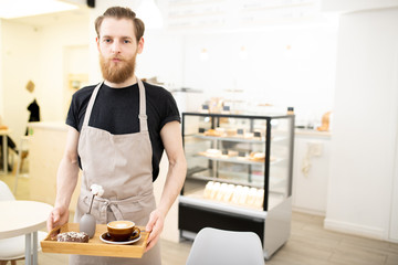 Serious handsome young barista man carrying tray with cappuccino and chocolate doughnut in modern...