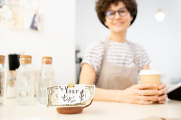 Close-up of tag Your tips and dollar attached to metal cup on counter, pretty lady with takeout coffee cup in background