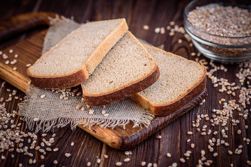 A loaf of rye bran bread with slices on sackcloth on cutting board and wheat on dark wooden background.