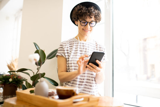 Smiling attractive modern young woman in hat and stripped tshirt sitting on window sill and listening to music in earphones while using smartphone in cafe