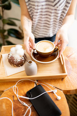Close-up of unrecognizable lady sitting at wooden table with tray and smartphone and drinking cappuccino in coffee shop