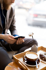 Close-up of wooden tray with morning cappuccino and chocolate doughnut, hipster man using smartphone in background