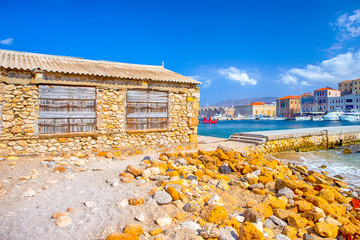 View of Chania old Venetian Port and Boats in Background. Lots of Stones on Sandy Beach in Front.
