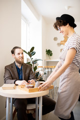 Smiling friendly waitress in hat bringing order to customer in modern coffee shop and putting tray on table, cheerful bearded man in earphones smiling at waitress