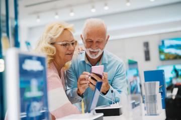 Mature couple leaning on stand and trying out smart phone in tech store.