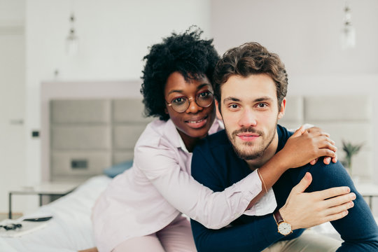 Happy African american girl embracing her handsome caucasian boyfriend while sitting on bed in bedroom with modern grey colored interior and big windows.