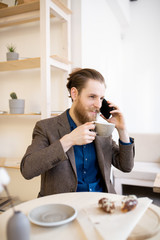 Smiling positive handsome hipster businessman with beard sitting at table and drinking coffee while communicating on mobile phone in cafe