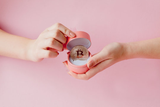 Woman's hands holding bitcoin in pink gift box on a pink background, symbol of virtual money