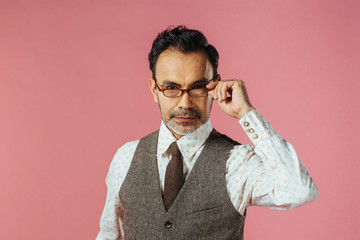 Portrait of a mature man in brown vest holding his glasses, isolated on pink studio background