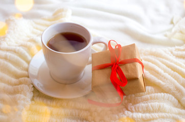 Fototapeta na wymiar A cup with tea or coffee and a box with a gift tied with a red ribbon on a white knitted background. Romantic breakfast in bed for Valentine's Day or a gift for mother's day 